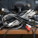 Collection of very tangled cords of all different types sitting behind an electronic screen on a dusty shelf.