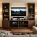 A high-angle photograph looking over the back of a beige sectional sofa into a modern, organized living room. The center of the room features a large walnut entertainment center with a wall-mounted TV displaying a mountain landscape, flanked by open bookshelves with decor, plants, and books. Below the TV is a soundbar, several charging electronics, and multiple game controllers. An open lower cabinet reveals a couple of gaming systems. A rustic wooden coffee table with books sits on an area rug on a hardwood floor. Two standing lamps provide warm ambient light.